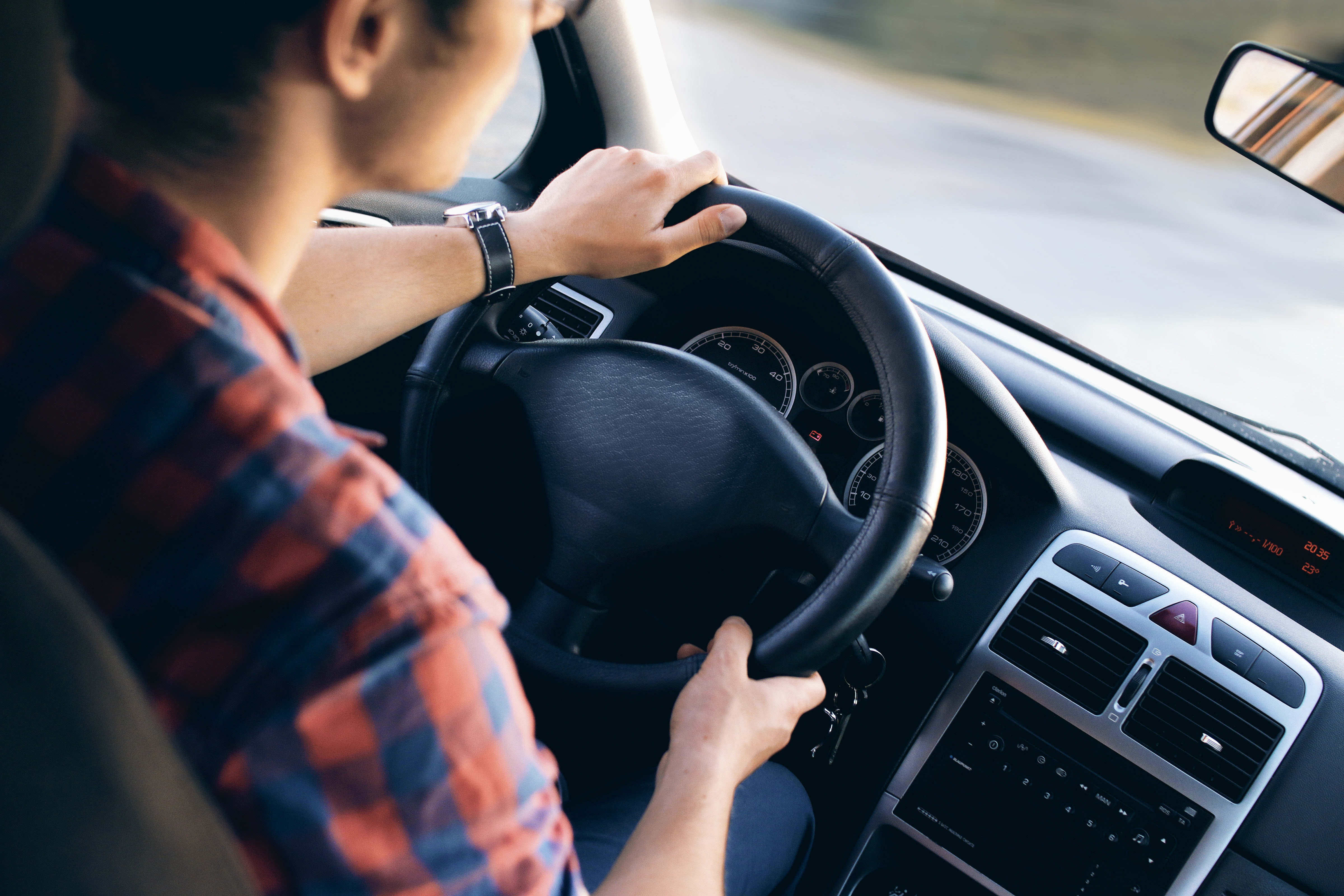 Imagem de dentro de um carro: a esquerda um motorista segurando o volante com as duas mÃ£os, a direita o painel do carro e o retrovisor e o parabrisa
