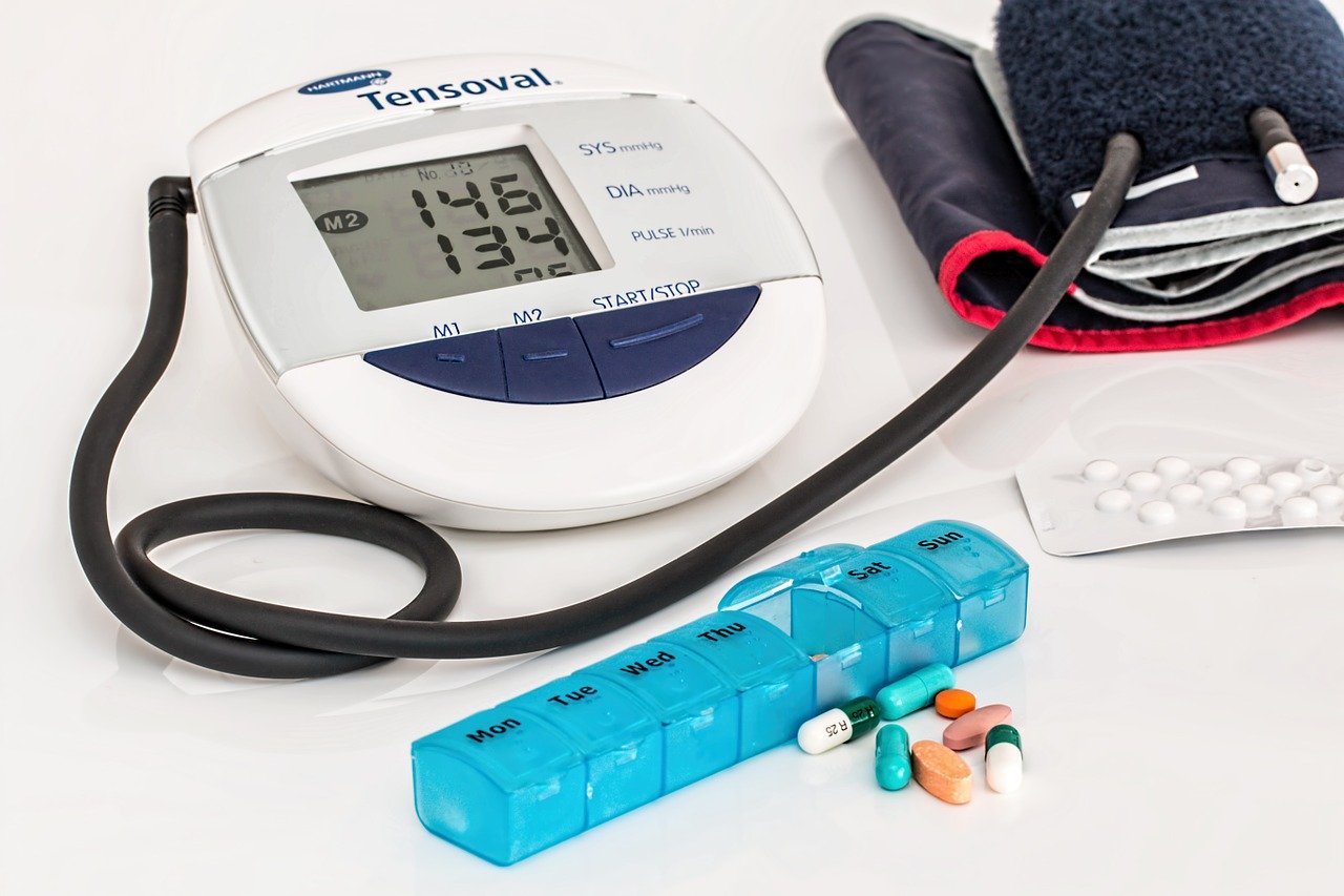 Foto de um medidor digital de pressão, de um porta medicamentos azul e de algumas cápsulas de remédios coloridas.