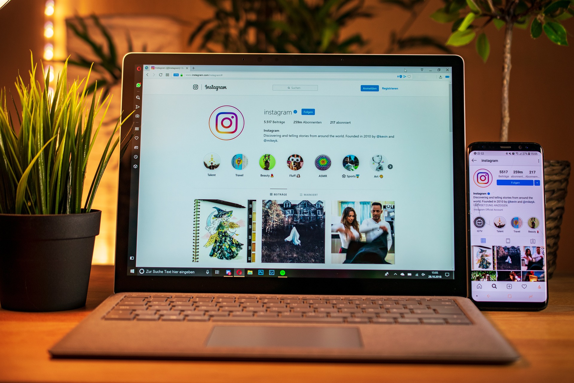 Imagem de uma mesa contendo um vaso de plantas à esquerda, um notebook ao centro e um celular à direita, ambos exibindo a tela do aplicativo instagram.